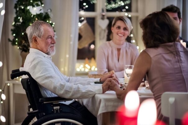 Elderly man in a wheelchair at the dinner table holding hands with his family during the holidays